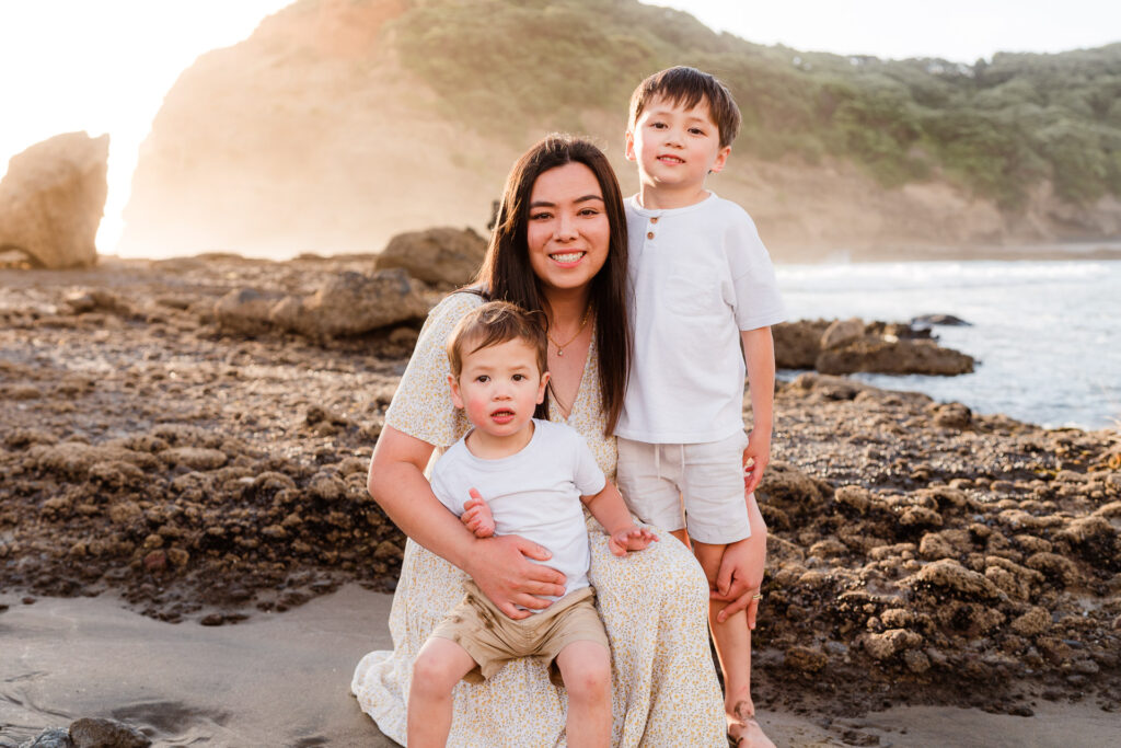Mum holding her son close with golden light surrounding them at Bethells Beach – intimate family portrait by West Auckland photographers.