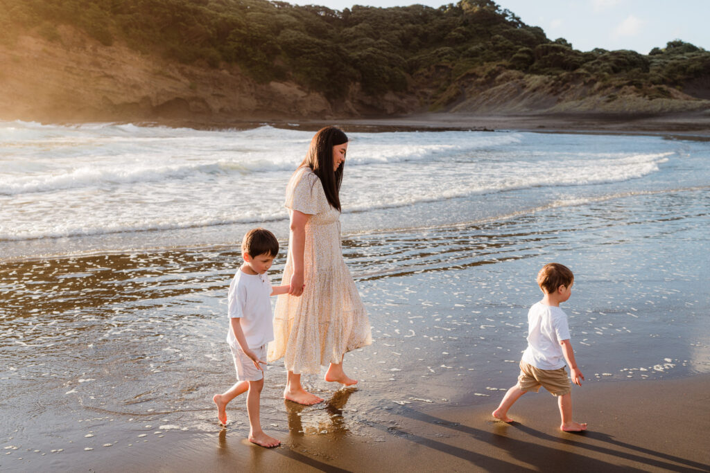 Mum walking with two young boys on Bethells Beach during a golden hour family shoot – Auckland family photographers capturing natural moments.