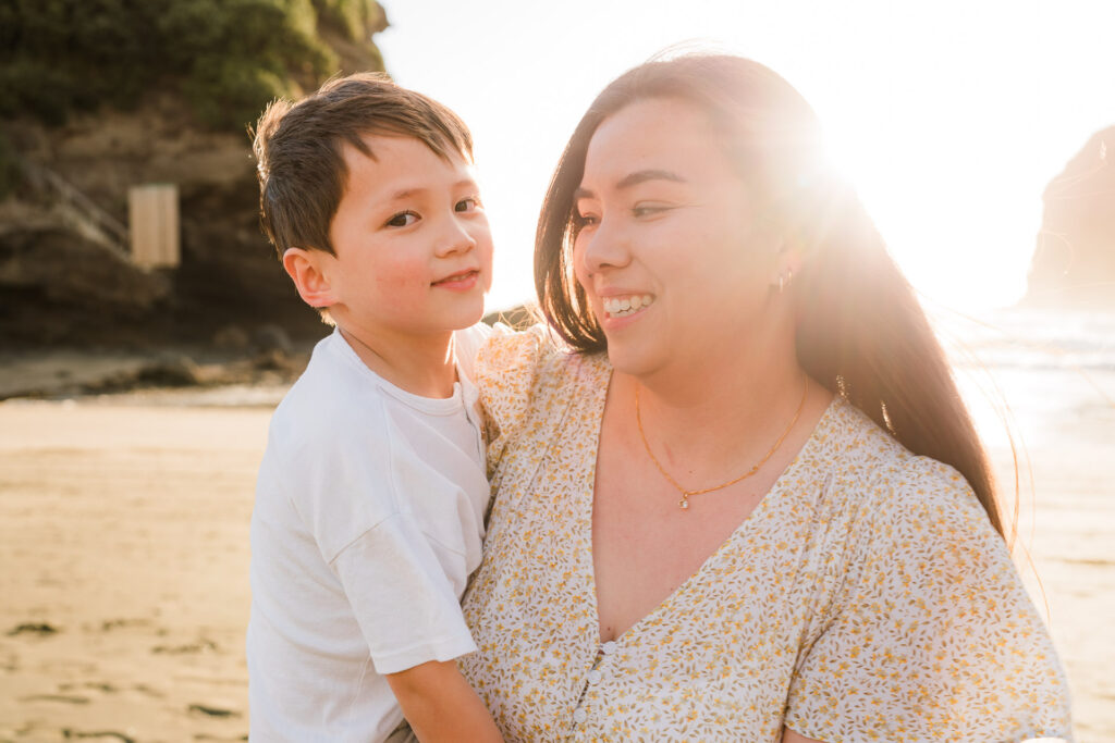 Young boy cuddling his mum on Bethells Beach in golden light – genuine family connection captured by Auckland photographers.