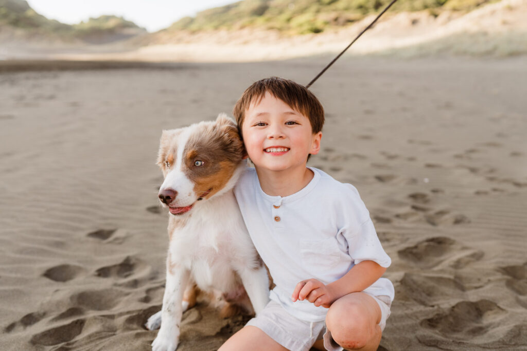 Young boy cuddling family dog on Bethells Beach during a sunset family session – authentic children’s photography Auckland.