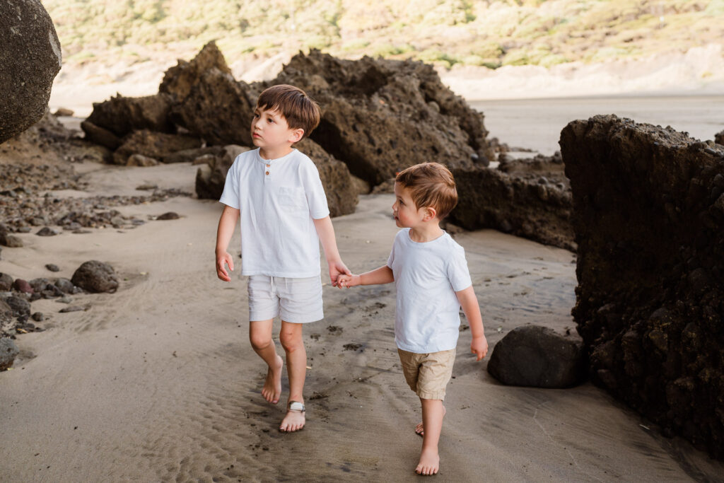 Two boys walking hand-in-hand along the shoreline at Bethells Beach – sibling connection captured by Auckland family photographers.
