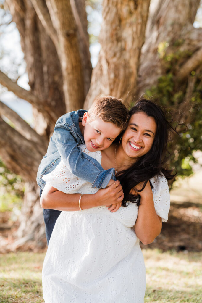 Mother and son laughing and cuddling by the water’s edge at Cornwallis Beach – West Auckland family photographers capturing connection.