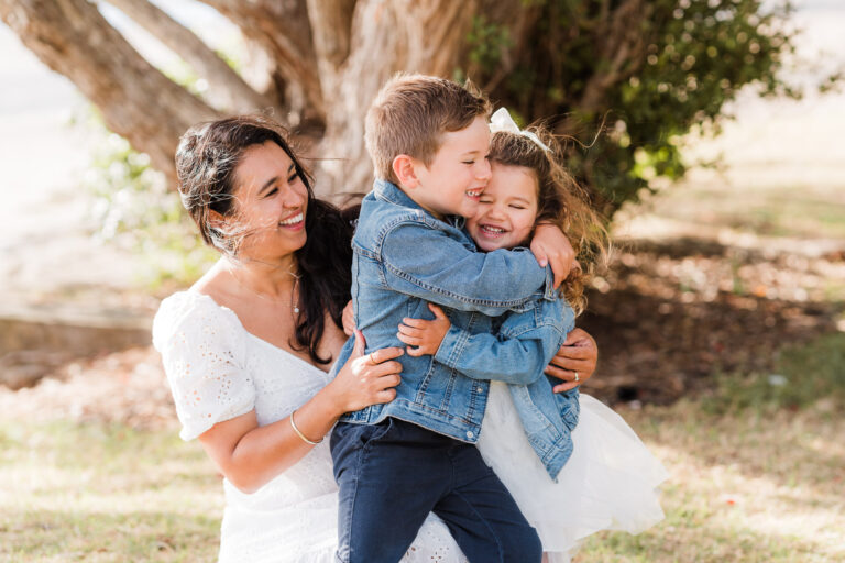 Mum holding baby daughter as older brother cuddles her during a family shoot on the beach – authentic family moments by Auckland family photographers.