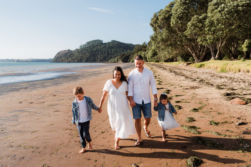 Family of four walking hand-in-hand along Cornwallis Beach during a relaxed sunset shoot – captured by Auckland family photographers.