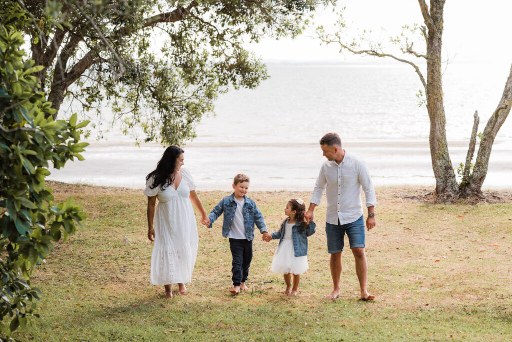 Joyful moment as parents and kids walk hand-in-hand and laugh together at Cornwallis Beach – outdoor family shoot by Auckland photographers.