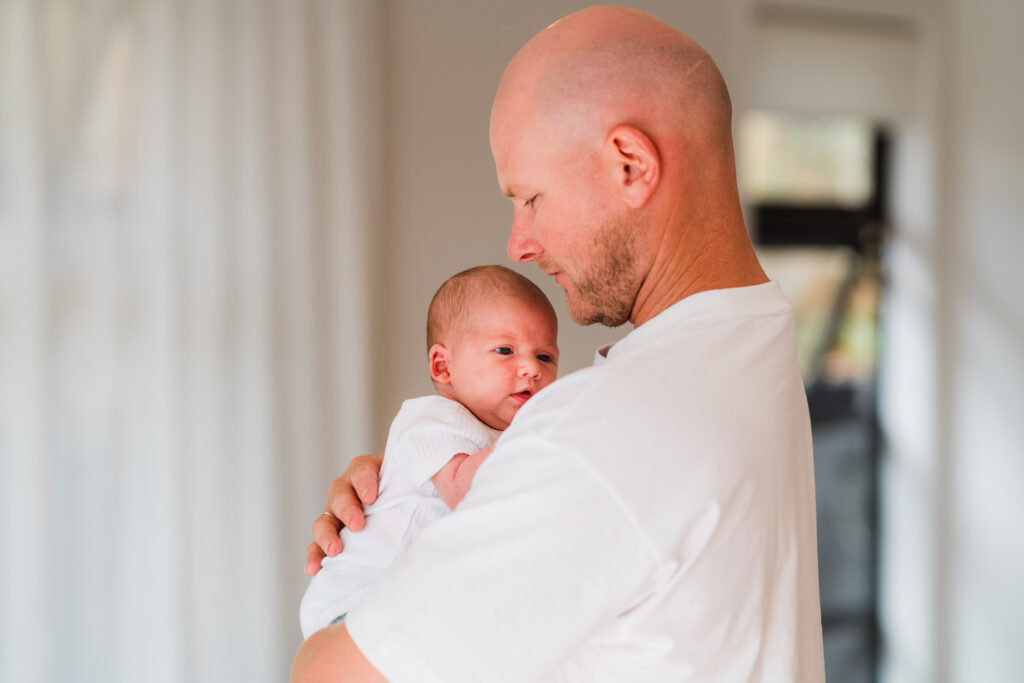 Newborn baby cuddled in dad’s arms during a lifestyle home shoot in Auckland – authentic family photography by Auckland photographers.