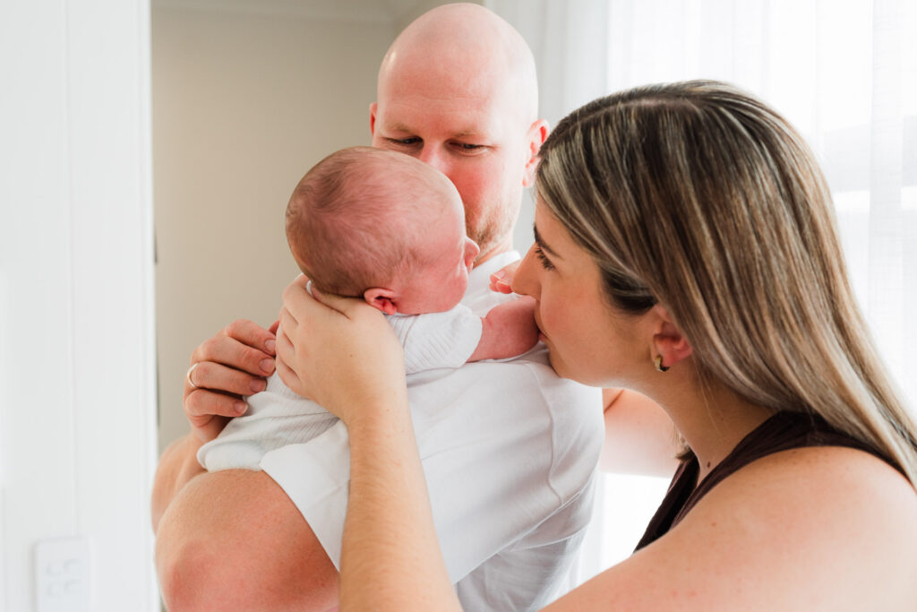 Family of three with newborn resting on mum’s shoulder as she kisses the baby – lifestyle newborn photography Auckland.