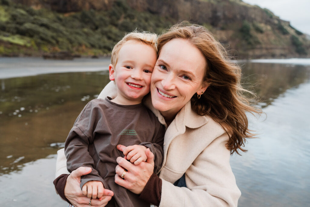 Mum hugging her son at the shoreline during a golden hour mini session at Muriwai Beach in Auckland.
