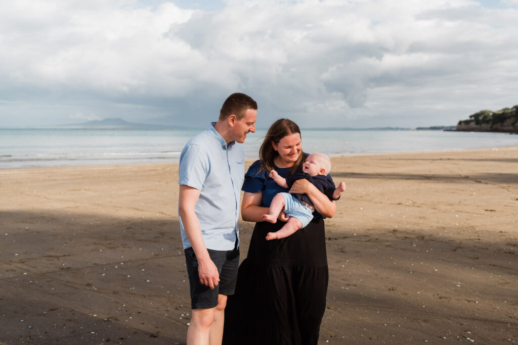Parents holding their baby together while walking along Waiake Beach in late afternoon light – natural family moments in North Shore Auckland.