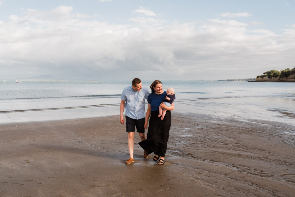 Family of three walking hand-in-hand along the shoreline at Waiake Beach – candid family session in Auckland.