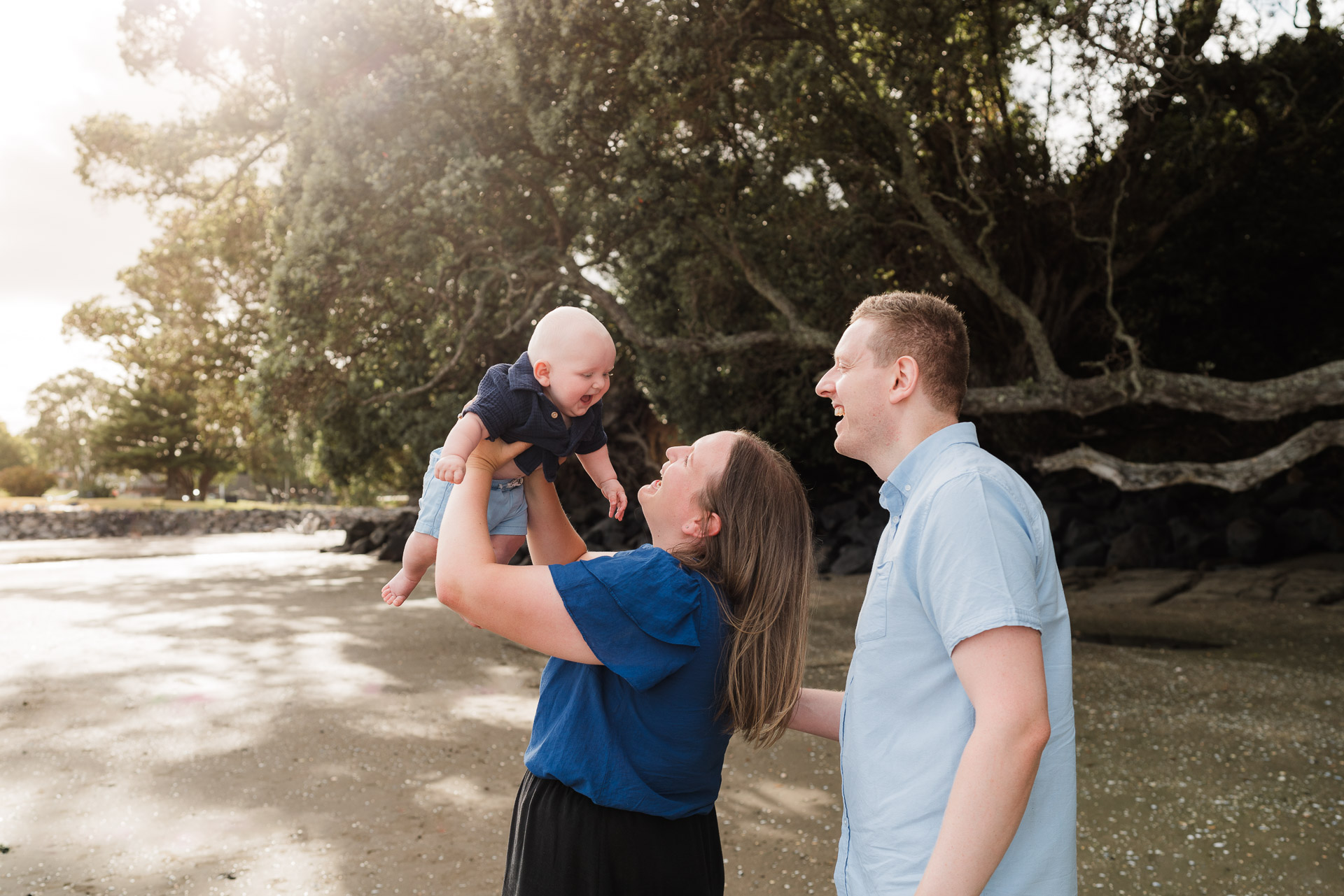 Baby cuddled close by dad near trees at Waiake Beach with warm golden light – intimate North Shore family photography.