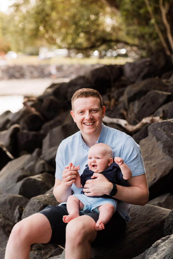 Baby sitting on dad’s knee on rocks at Waiake Beach – natural and warm family photography on Auckland’s North Shore.