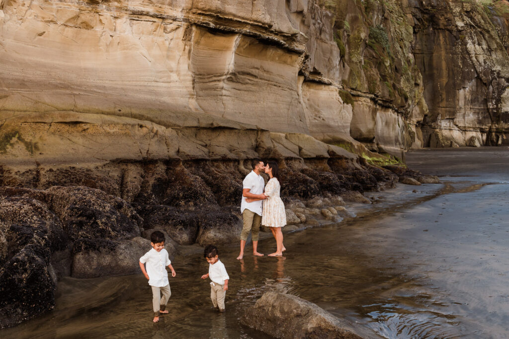 Candid moment of parents sharing a kiss while their children play in the sand, photographed at Maori Bay by Wonderferris.