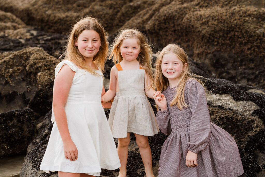 Three sisters hugging and smiling during a sunset mini session at Muriwai Beach, photographed by Wonderferris.