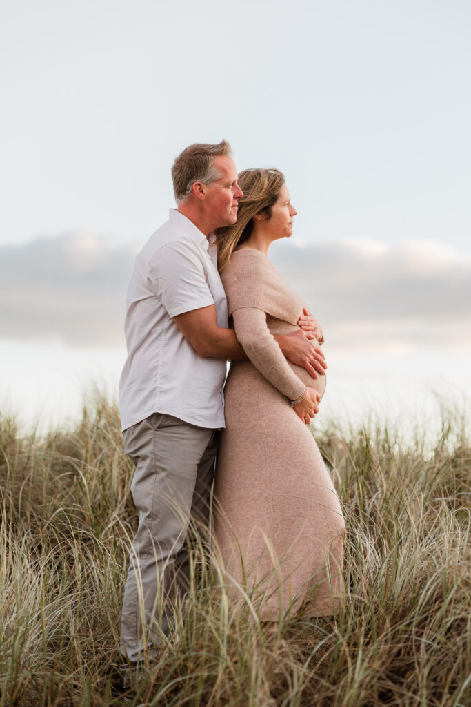 Side profile of couple embracing baby bump and gazing into the distance with soft sunrise light — emotive maternity photography by a West Auckland-based family photographer.