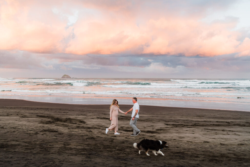 Wide shot of expecting couple walking hand in hand along Muriwai Beach with their dog at sunrise — natural, candid family photography by Auckland photographer based in Kumeu.