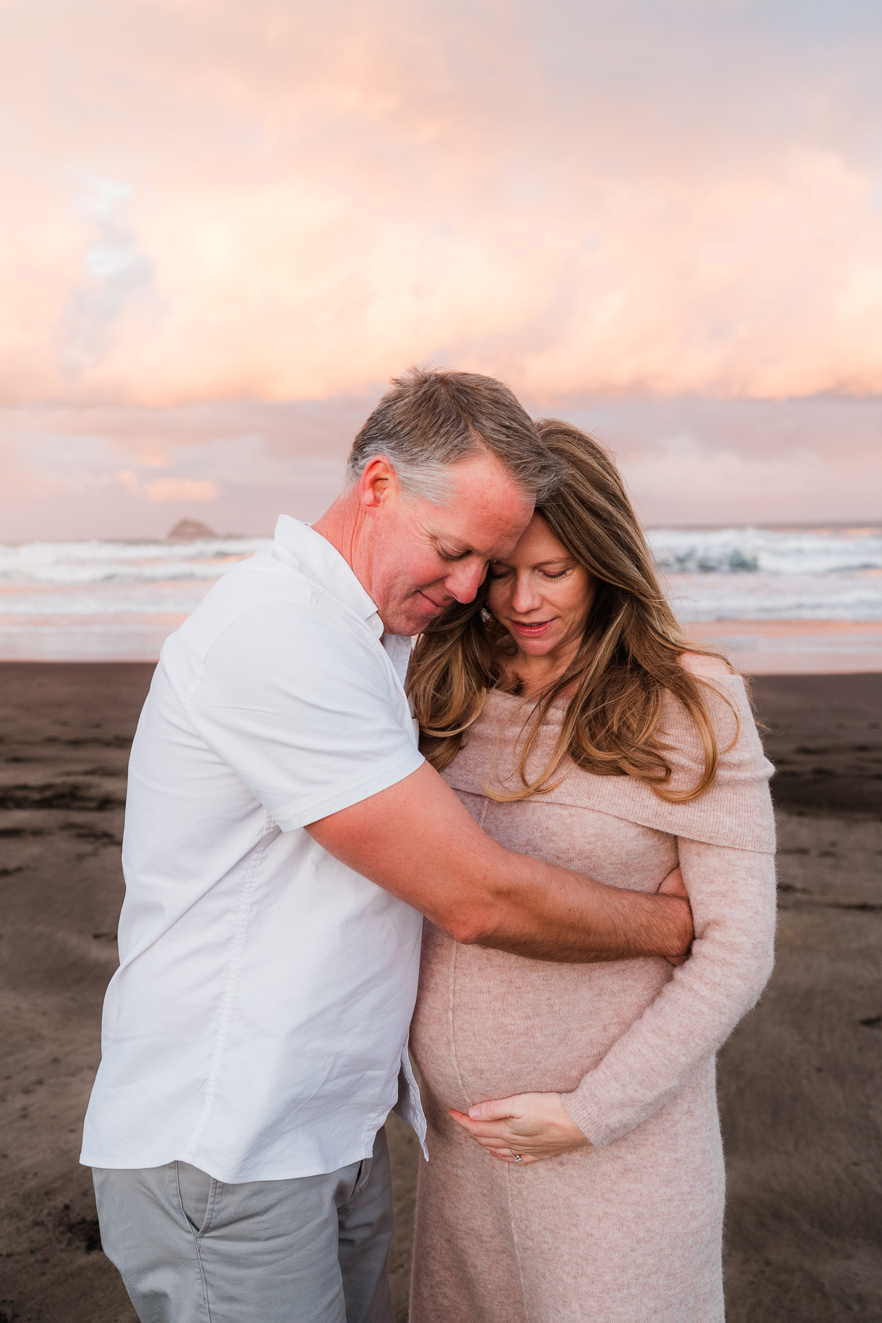 Expecting couple cuddling baby bump at sunrise on Muriwai Beach, with soft pastel skies — captured by Kumeu-based Auckland family photographer specialising in maternity and family sessions across West Auckland.