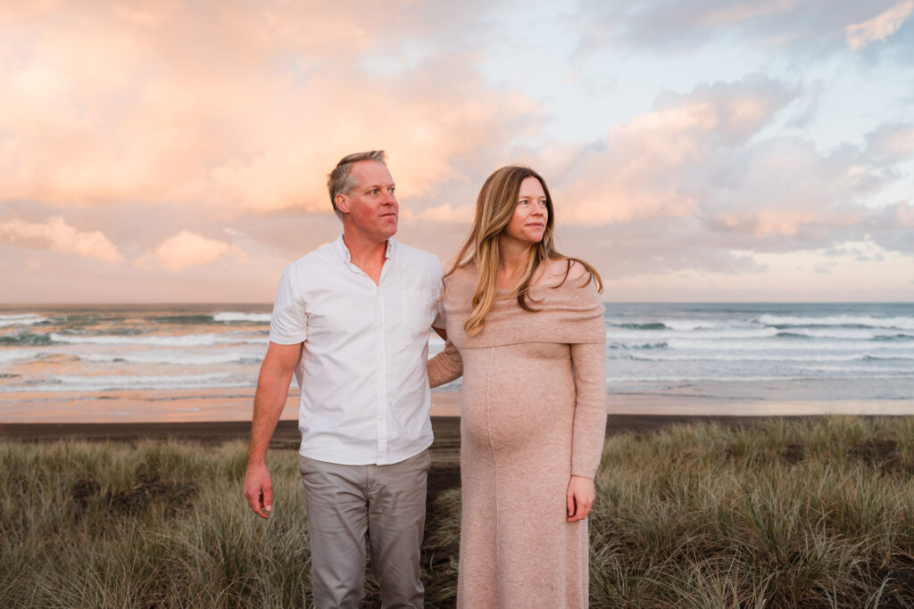 Couple looking out towards the ocean with baby bump in view and pastel sunrise skies behind — peaceful West Auckland maternity portrait by a family photographer in Kumeu.