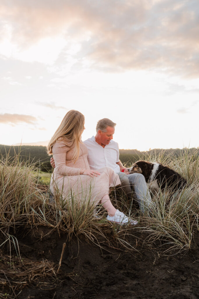 Expecting couple sitting in the sand dunes at Muriwai Beach with their dog — relaxed, lifestyle maternity session by Auckland family photographer in Kumeu.