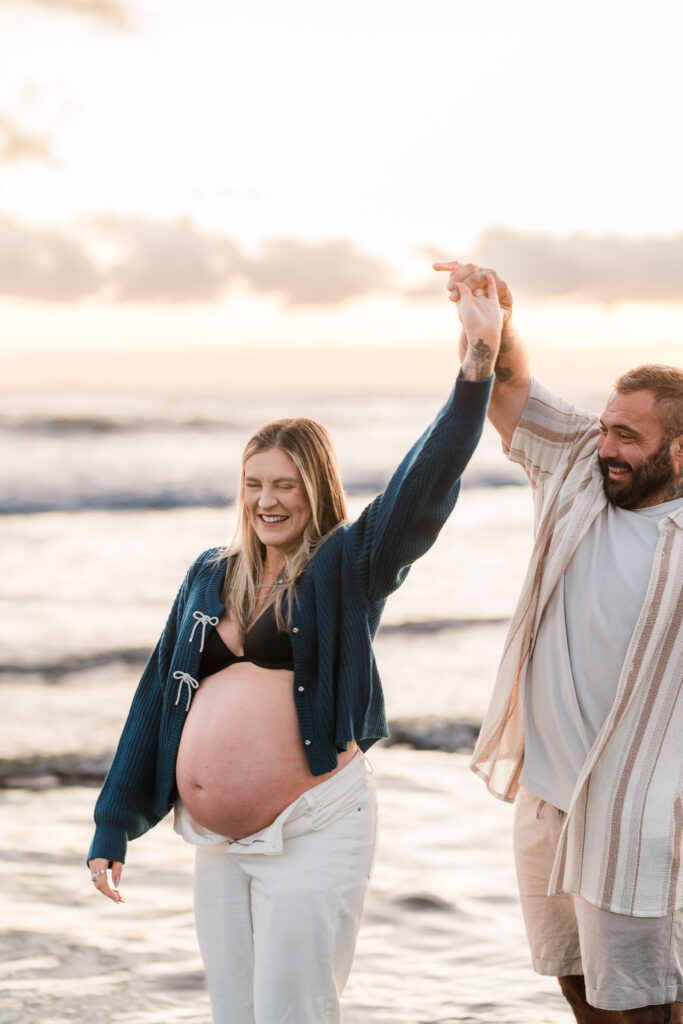 sunset muriwai maternity shoot of mum to be in denim jacket twirling and dancing with partner