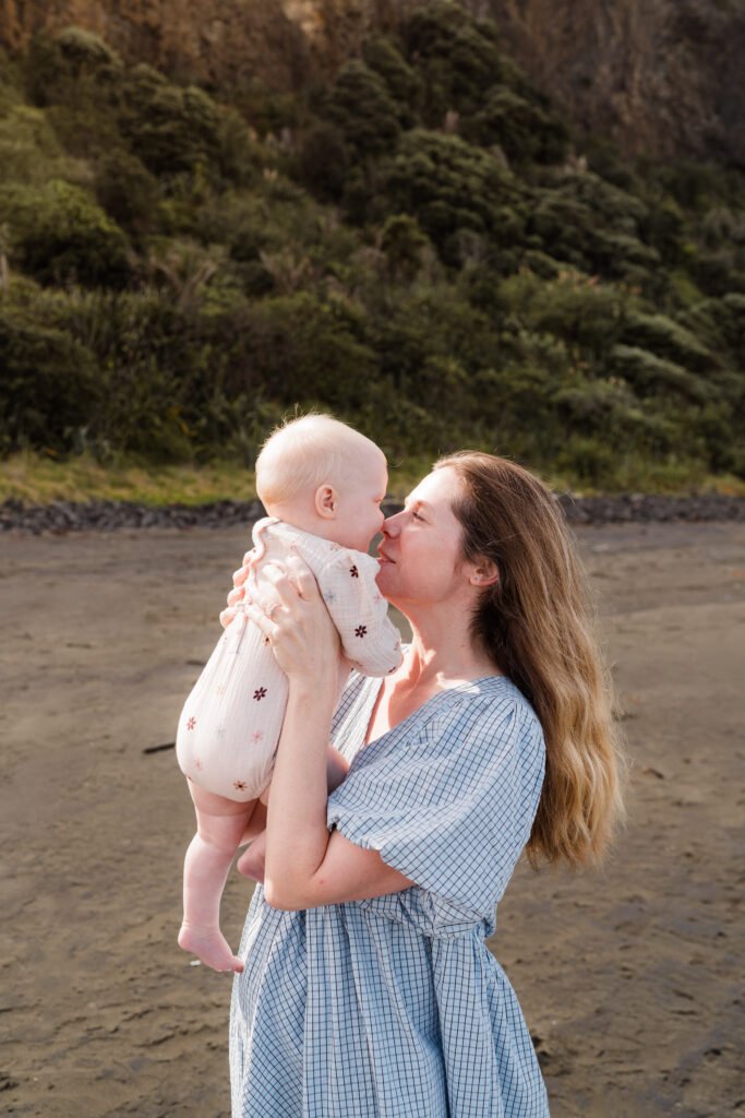 A woman holding a baby up near a sandy beach with a green hillside in the background.