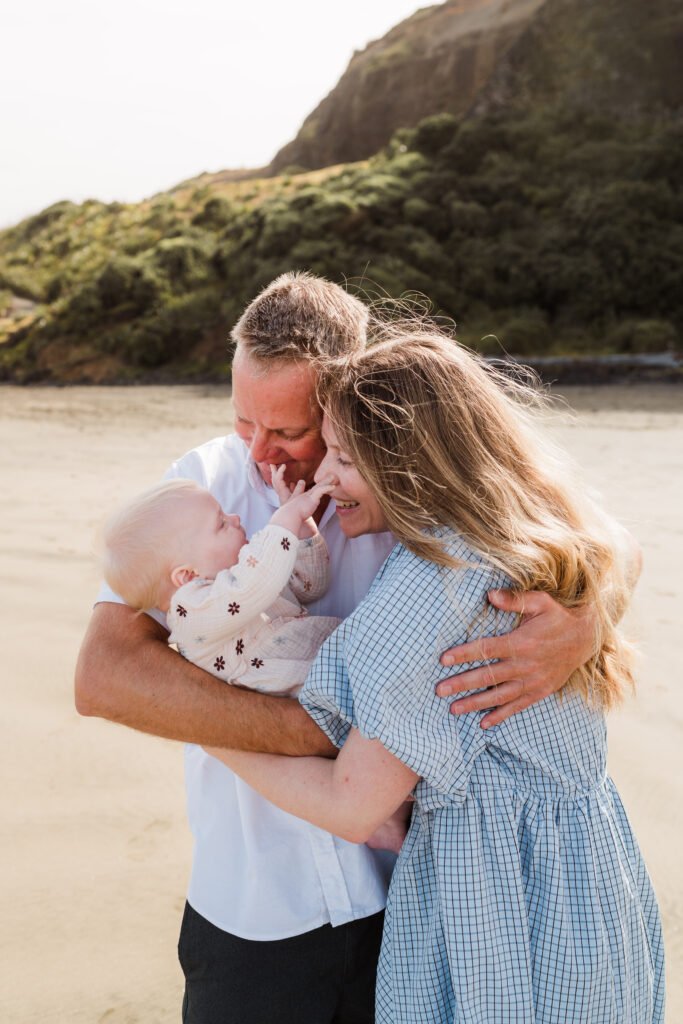 Couple holding and cuddling a baby on a sunny beach with a rocky hill in the background.
