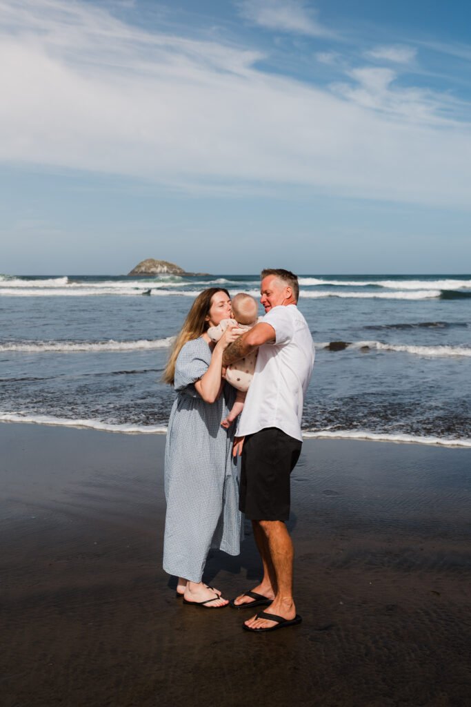 A couple holding their baby on a beach, standing in shallow tidal water as waves roll in, with a small island in the distance.