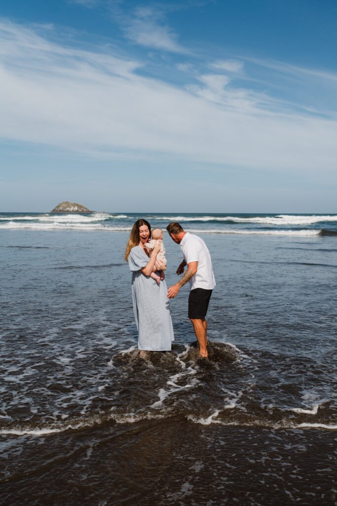 A woman holding a baby on a rocky beach while a man stands beside them as waves wash over the shore.