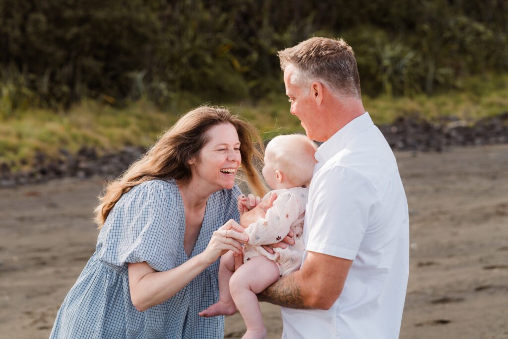 Couple smiling and holding their baby on a sandy beach near calm water and greenery in the background.