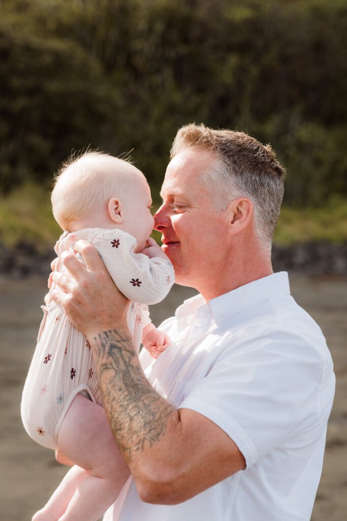 Father holds baby close, noses nearly touching, outdoors by water and greenery.