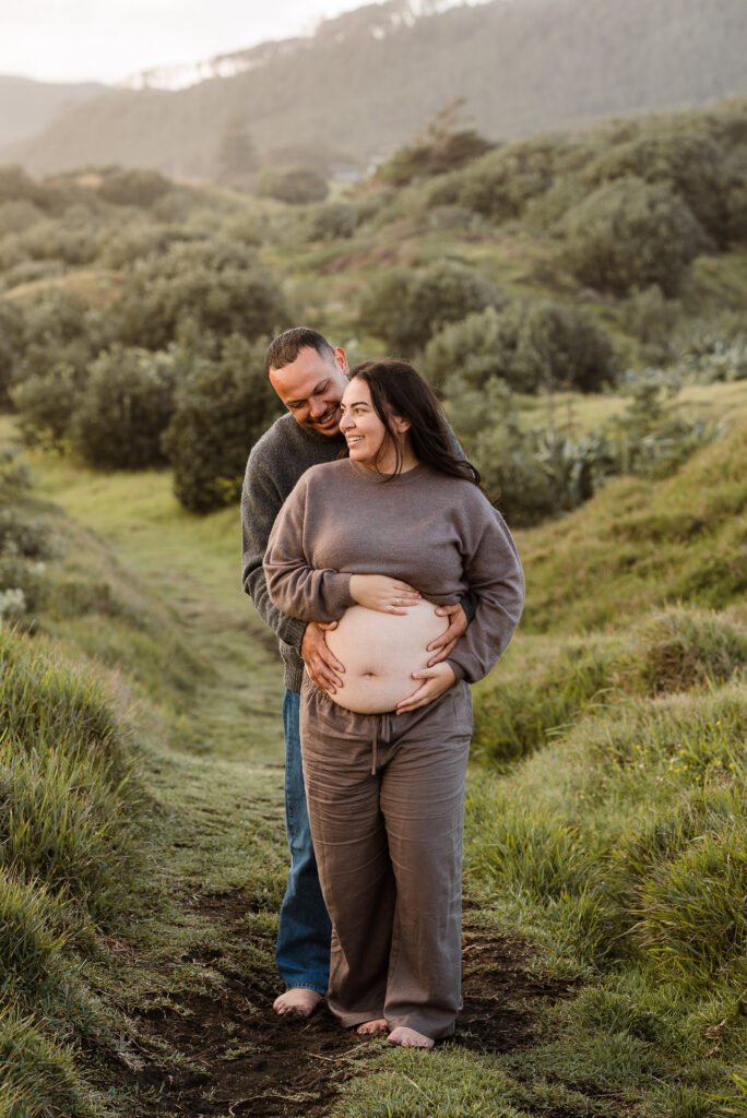 Couple embracing in a grassy field, woman cradles her pregnant belly, both smiling warmly, surrounded by lush greenery.