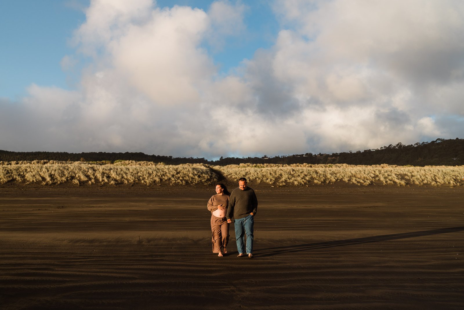 Pregnant woman and partner walk barefoot on a dark-sand beach, with tall dunes and a cloudy sky behind them.