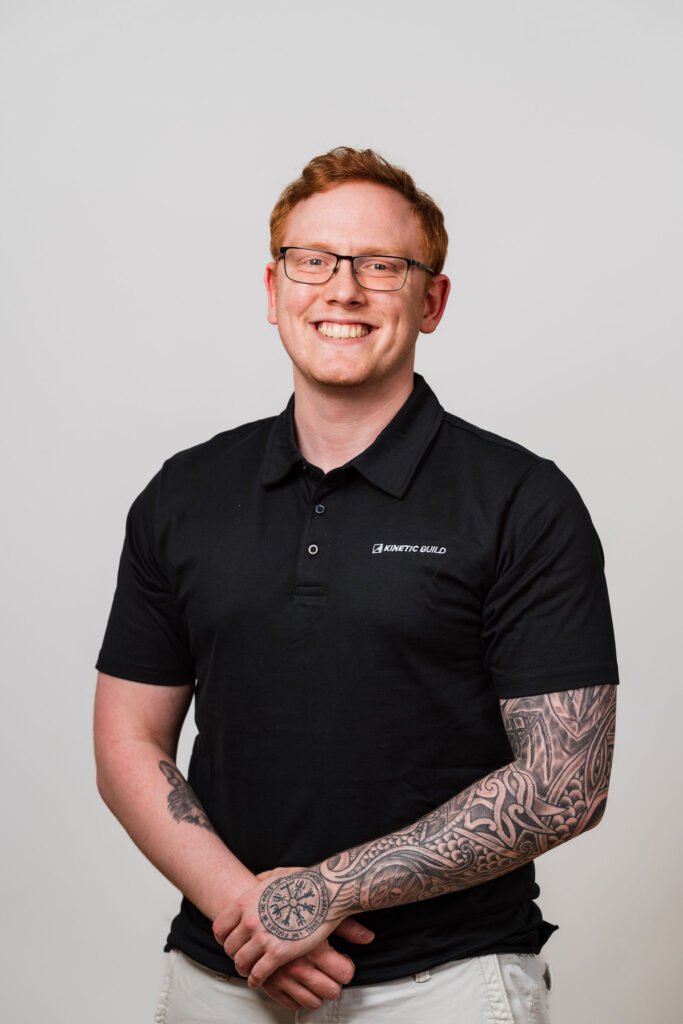 Smiling person with glasses and arm tattoos, wearing a black polo shirt with "Kinetic Build" logo, stands against a plain background.