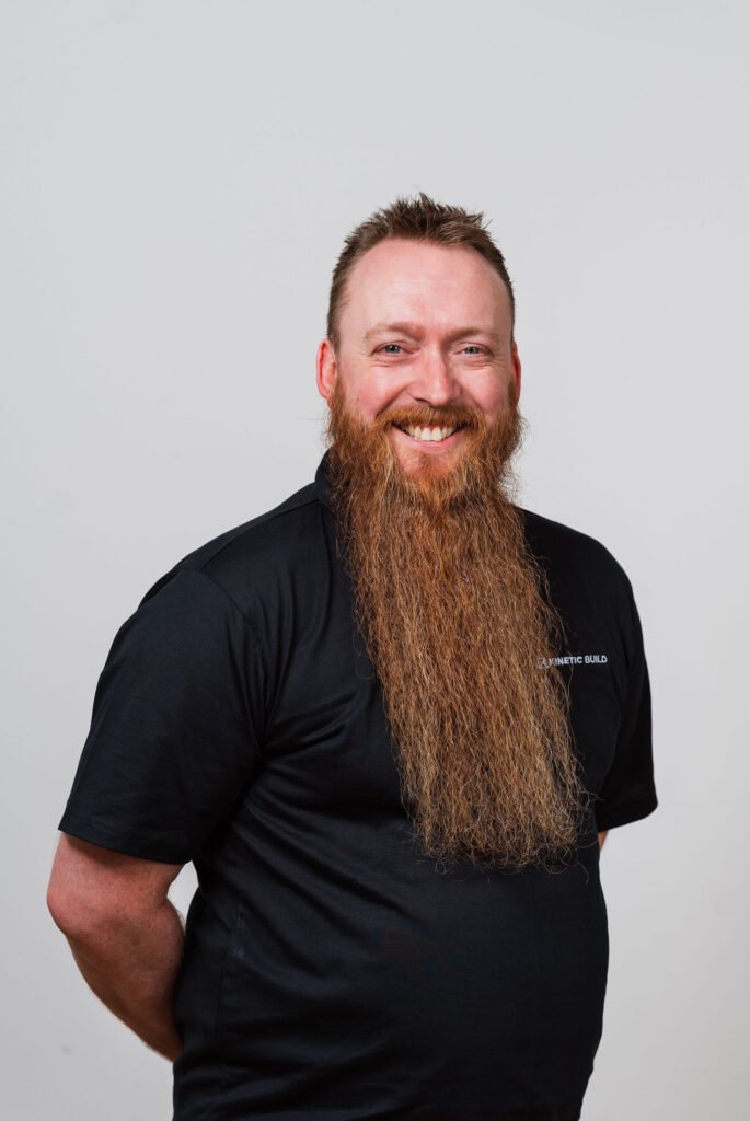 Smiling man with a long beard wearing a black shirt stands against a plain white background.