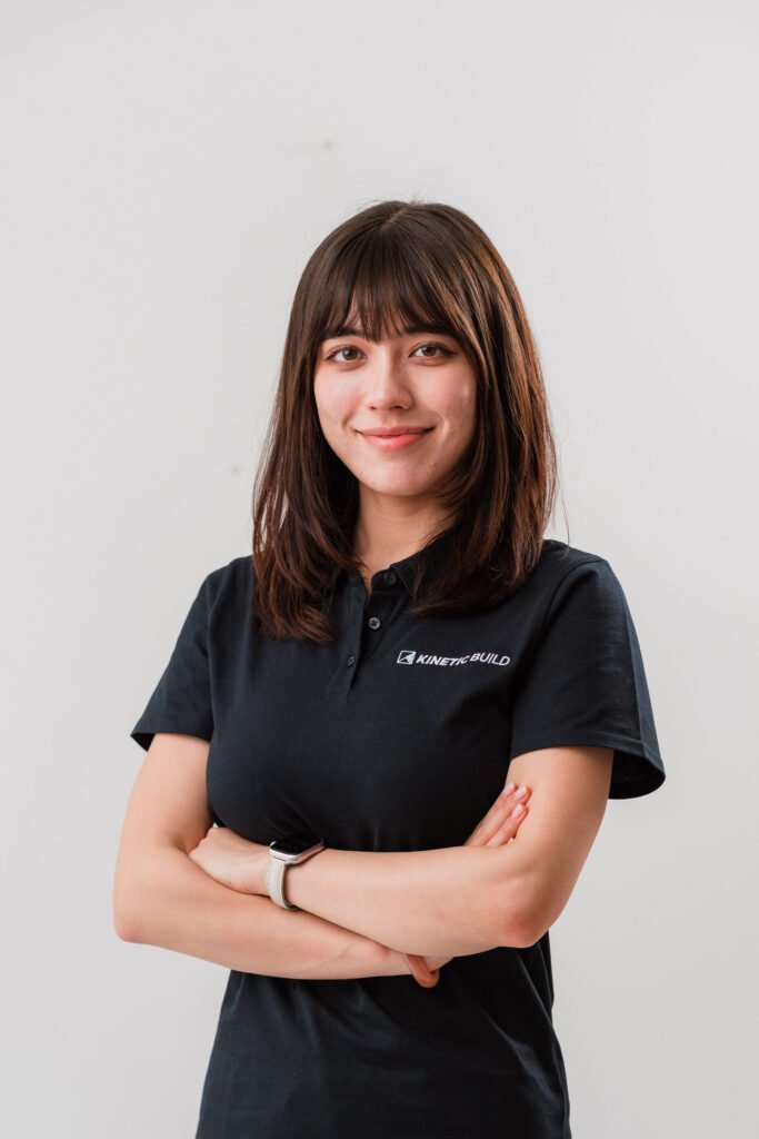 Smiling woman with brown hair, wearing a black "Kinetic Build" polo, stands with arms crossed against a plain background.