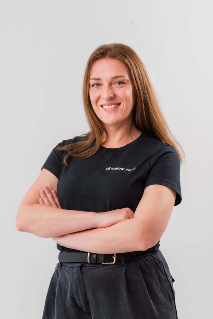 Smiling woman with long brown hair, wearing a black t-shirt and pants, stands with arms crossed against a plain white background.