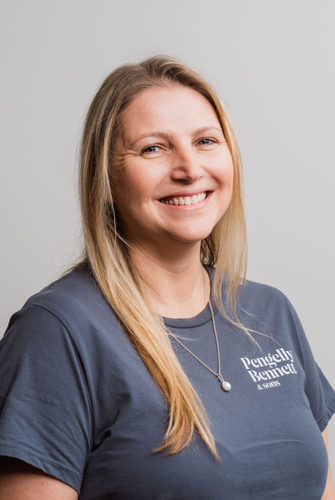 Smiling woman with long blonde hair in a gray "Pengelly Bennett & Sons" shirt, wearing a necklace, against a plain background.