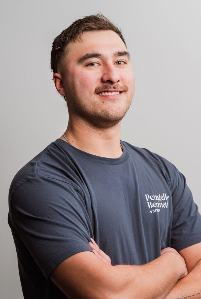 Smiling man with short hair and a mustache, wearing a dark blue "Pengelly Bennett & Sons" shirt, arms crossed, against a gray background.