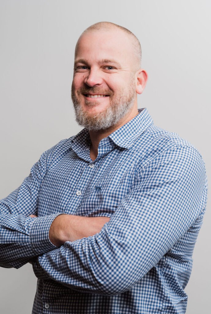 Smiling man with a beard and shaved head, wearing a blue checkered shirt, arms crossed against a plain background.